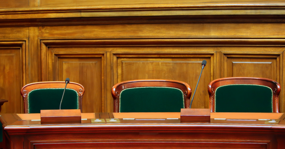 Image shows the wooden bench which is a mahogany table with three padded chairs for three judges in court